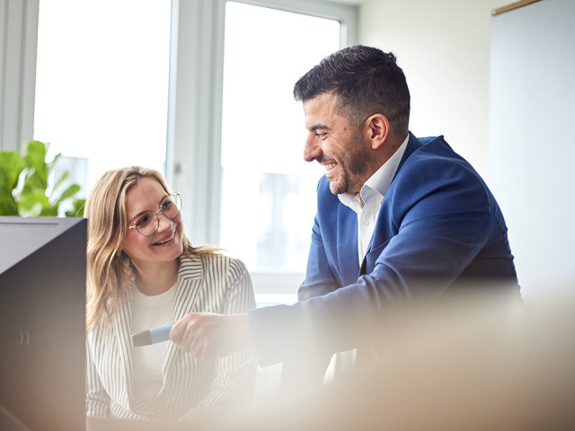 Zwei TriFinance-Consultants besprechen l&auml;chelnd etwas vor einem Computer-Bildschirm, symbolisch f&uuml;r operative Entlastung und Team-Support im Accounting.