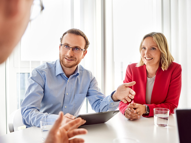 Ein Berater im Hemd sitzt neben einer Kollegin in rotem Blazer am Tisch und pr&auml;sentiert einem Gegen&uuml;ber Analysen auf einem Tablet, symbolisch f&uuml;r modernes Controlling, Business Intelligence und datenbasierte Entscheidungen.