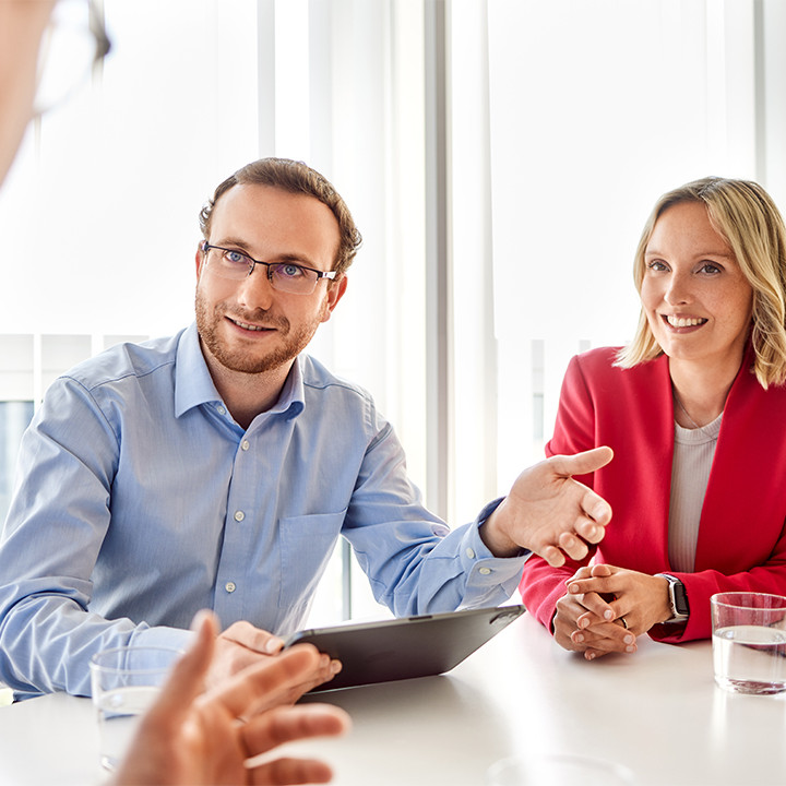 Ein Berater im Hemd sitzt neben einer Kollegin in rotem Blazer am Tisch und pr&auml;sentiert einem Gegen&uuml;ber Analysen auf einem Tablet, symbolisch f&uuml;r modernes Controlling, Business Intelligence und datenbasierte Entscheidungen.
